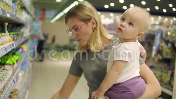 漂亮的女人戴着眼镜抱着她的孩子一边在超市的货架上挑选婴儿食品视频的预览图
