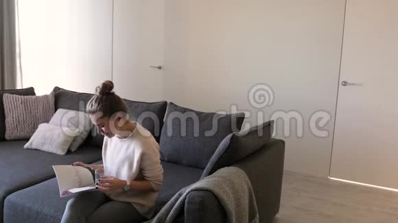 年轻女子在客厅里拿着一杯水看杂志一个女人在家休息看杂志视频的预览图