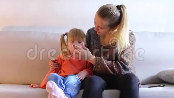 年轻的母亲和女儿坐在沙发上擦拭婴儿的鼻子视频的预览图