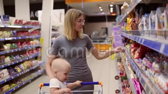 漂亮的年轻妈妈正在超市里挑选糖果和蛋糕而她的小宝宝正坐在杂货店里视频的预览图