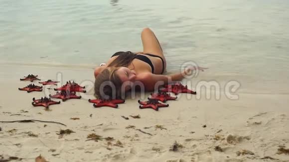 一个年轻美丽的女人躺在海滩上被红色的星星鱼包围着视频的预览图