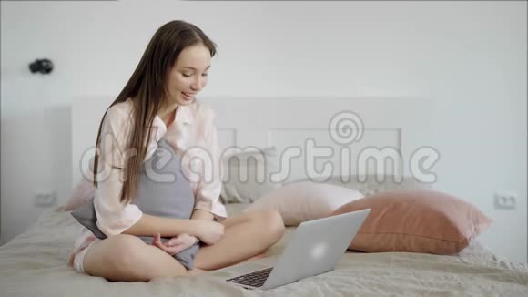 漂亮的黑发女孩正在使用笔记本电脑室内坐在床上视频的预览图