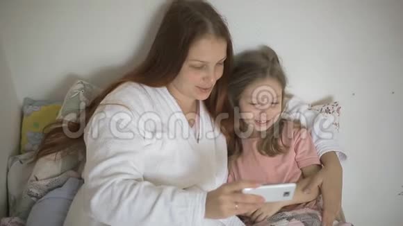 一个小女孩和妈妈躺在床上看着智能手机屏幕上的卡通片儿童和数字视频的预览图