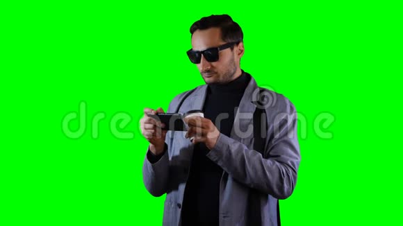 穿着西装和黑色太阳镜的人在手机上输入一条信息然后拿着咖啡杯绿色屏幕录像视频的预览图