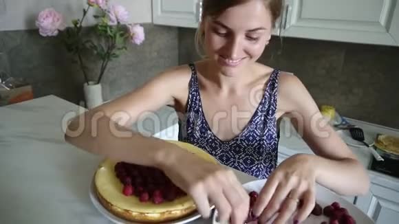 年轻微笑的女人在家里的厨房里用覆盆子装饰蛋糕的顶部奶酪蛋糕视频的预览图