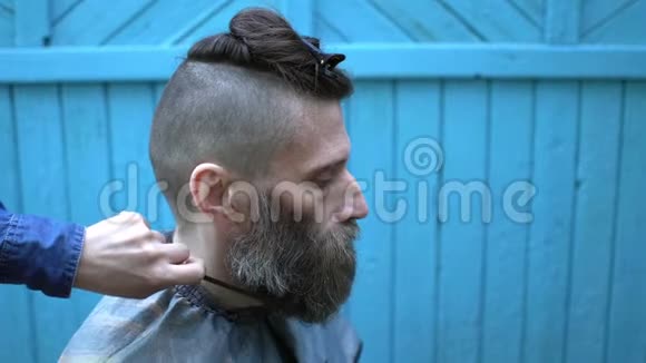 女理发师在实验性户外理发店修剪奇怪的中年嬉皮士的胡须男人看着视频的预览图