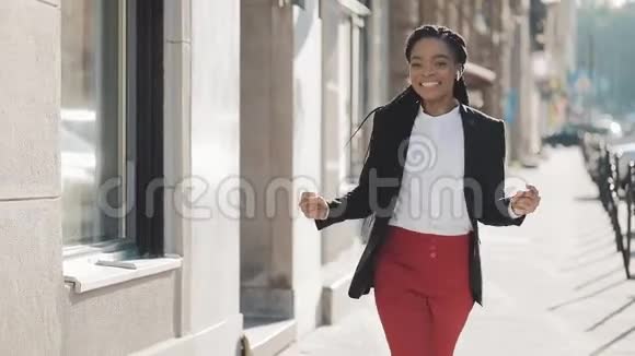 年轻快乐的非裔美国女商人戴着耳机在城市的大街上走着一边听着音乐视频的预览图