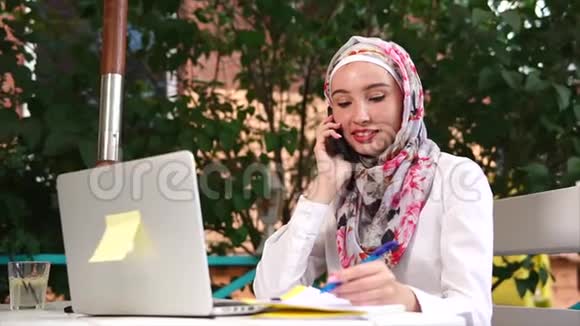 年轻的穆斯林妇女在手机上聊天在笔记本上做笔记视频的预览图