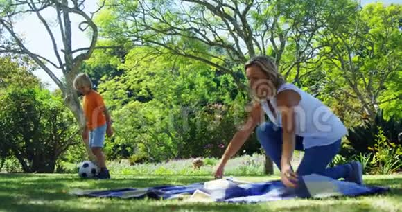 妈妈在公园铺野餐毯儿子在公园踢足球视频的预览图