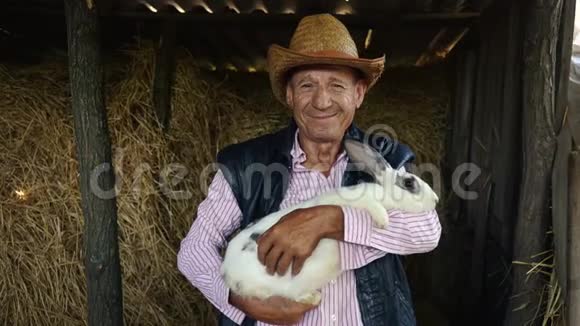 一个戴草帽的老农正在抱一只大白兔干草背景下男人的肖像视频的预览图