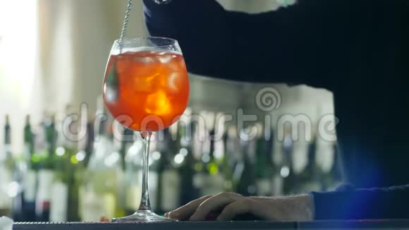 手里拿着长长的金属勺子的酒吧老板用冰块和杯子里的酒做明亮的鸡尾酒视频的预览图