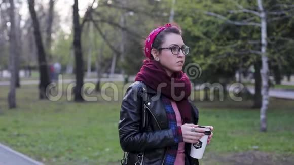 美丽年轻时尚的女人带着一杯咖啡在公园里散步穿皮夹克的现代女孩笼子里的衬衫视频的预览图