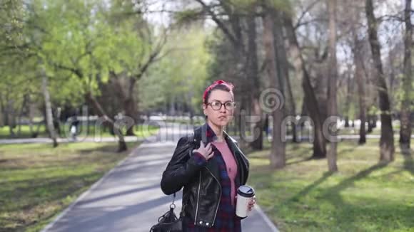 美丽年轻时尚的女人带着一杯咖啡在公园里散步穿皮夹克的现代女孩笼子里的衬衫视频的预览图