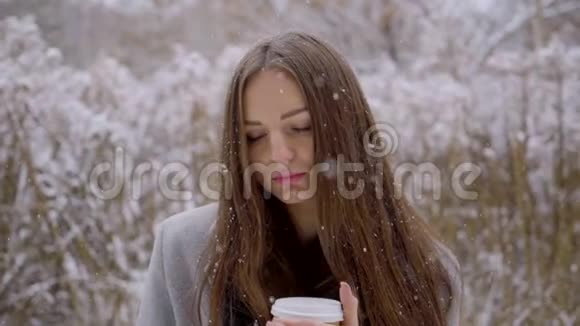 在冬天的公园里年轻美丽的悲伤女人端着一次性咖啡或茶杯坐在外面的长凳上视频的预览图