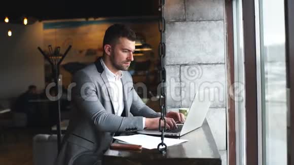 年轻英俊有魅力的胡子商人穿着西装坐在桌子旁在笔记本电脑上用手机打字视频的预览图