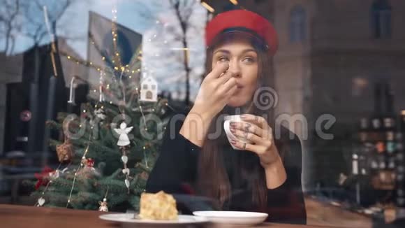 从外面看坐在舒适的咖啡馆里戴着红帽子喝着咖啡的迷人女人视频的预览图