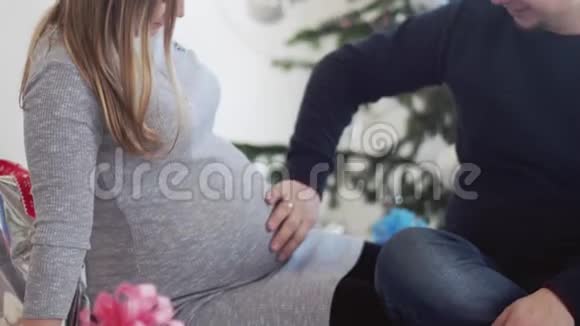 圣诞老人帽里快乐的年轻家庭丈夫摸着肚子他怀孕的妻子靠近美丽的装饰圣诞树慢慢慢慢视频的预览图