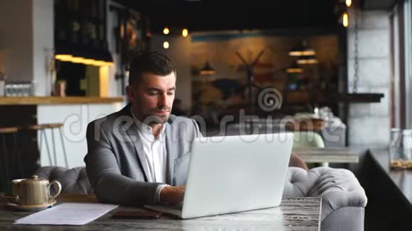 年轻的大胡子商人穿着灰色西装坐在咖啡馆的桌子上用笔记本电脑视频的预览图