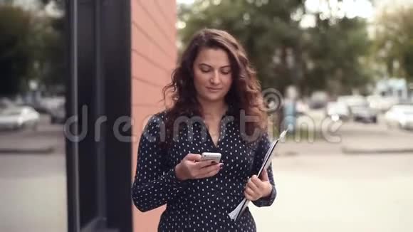 年轻漂亮的女商人走在办公楼旁边一边拿着文件一边打电话一边分心视频的预览图