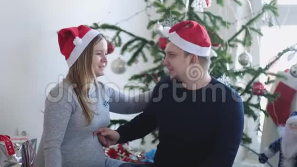 圣诞老人帽里快乐的年轻家庭丈夫在美丽的圣诞树旁拥抱他怀孕的妻子慢动作视频的预览图