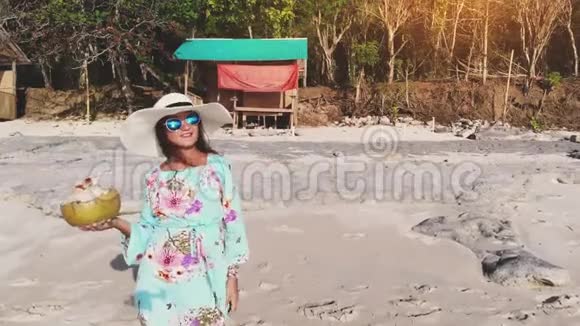 年轻漂亮的黑发女郎微笑着戴着帽子和太阳镜的女人放松在蓝色的海滩上喝椰子水视频的预览图