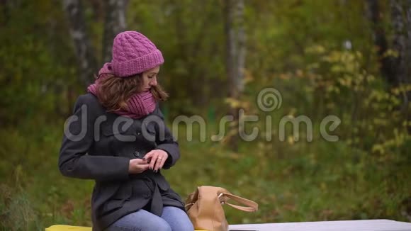年轻漂亮的女人从包里拿着手机坐在秋天公园的长凳上女人用智能手机视频的预览图