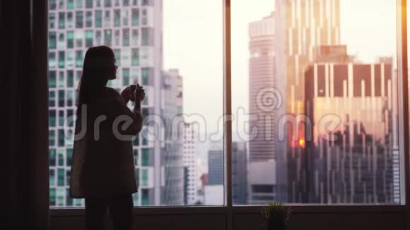 在美丽的日落中靠窗喝咖啡的美丽的女人站着看起来令人惊叹的城市景观慢动作视频的预览图