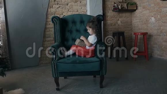 可爱的女孩打开礼物盒坐在客厅的椅子上带着圣诞树视频的预览图