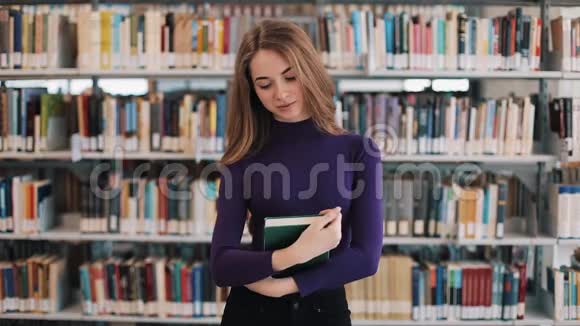 微笑的女学生拿着一本书站在图书馆的书架前视频的预览图