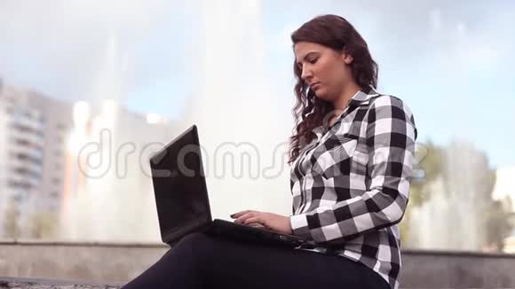 年轻漂亮的快乐女孩坐在城市的喷泉旁用笔记本电脑休息视频的预览图