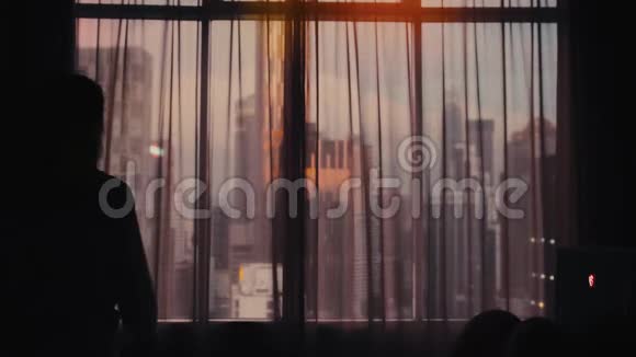 年轻女子打开大窗户上的窗帘伸出双手望着城市建筑上的公寓视频的预览图