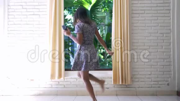 年轻有趣的漂亮黑发女人穿着灰色连衣裙手里拿着手机在窗边跳舞慢动作视频的预览图