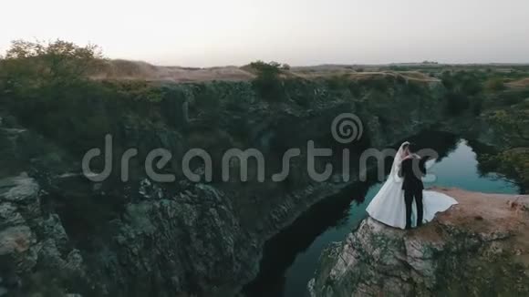 美丽的年轻新娘和新郎站在河底的悬崖上轻轻地拥抱在一起阳光明媚视频的预览图