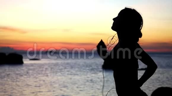 运动年轻女子欣赏日落的剪影带着耳机在海滩上奔跑慢动作1920x1080视频的预览图
