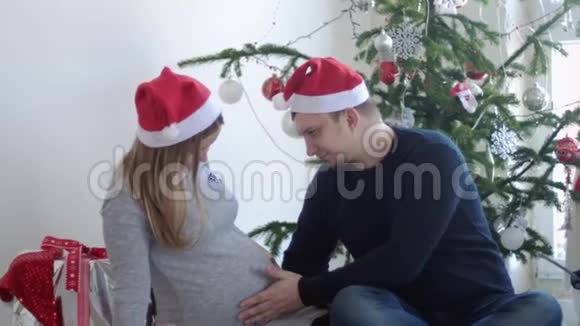 圣诞老人帽里快乐的年轻家庭丈夫摸着肚子他怀孕的妻子靠近美丽的装饰圣诞树慢慢慢慢视频的预览图