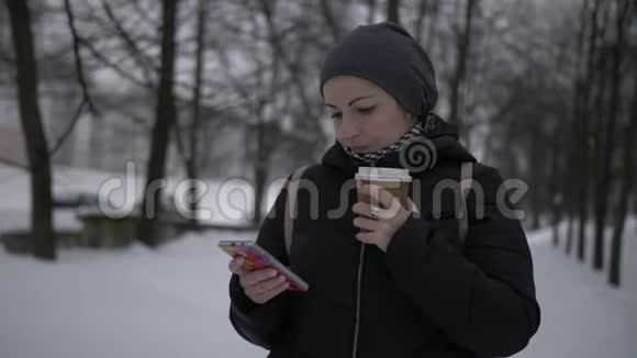 冬天雪上美丽的黑发女人喝咖啡用手机视频的预览图