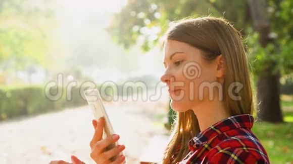 快乐的微笑年轻女人享受大自然坐在绿色公园的长椅上用手机说话视频的预览图