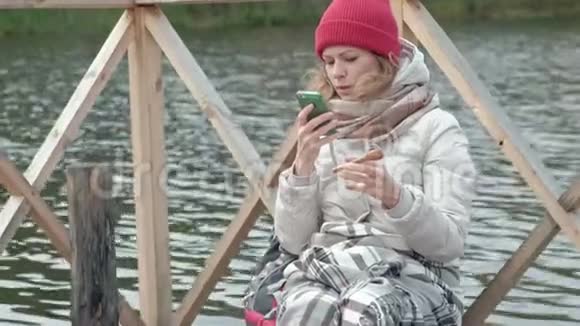 一位女游客穿着温暖的衣服在河岸附近的一座桥上背着一个背包上面铺着地毯喝着热茶视频的预览图