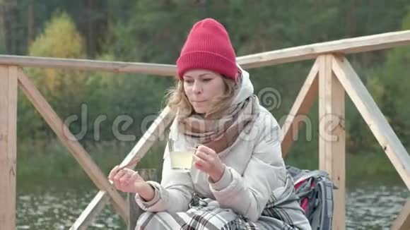 一位穿着温暖衣服的女游客在河岸附近的桥上背着一个背包上面盖着地毯喝着热茶视频的预览图