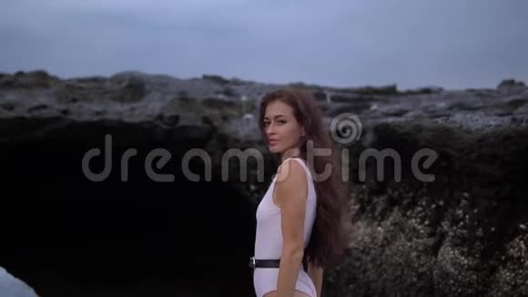 年轻漂亮的女性长着深色卷发身穿白色泳装在海滩上的岩石边散步视频的预览图