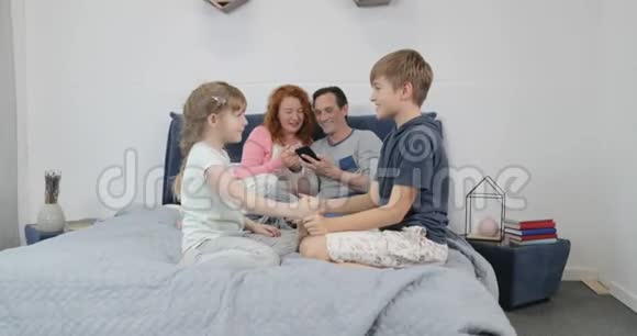 弟弟妹妹在父母的床上玩耍而母亲和父亲则使用手机家庭的早晨观念视频的预览图