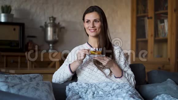 美丽的女人坐在沙发上裹着毯子喝茶视频的预览图