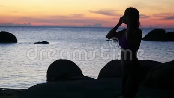 年轻女子欣赏日落的剪影带着耳机在海滩上奔跑慢动作1920x1080视频的预览图