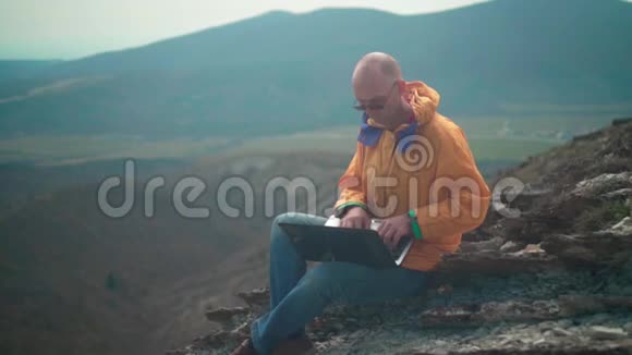 一个穿着黄色夹克蓝色牛仔裤和眼镜的人坐在悬崖边上在笔记本电脑上工作视频的预览图