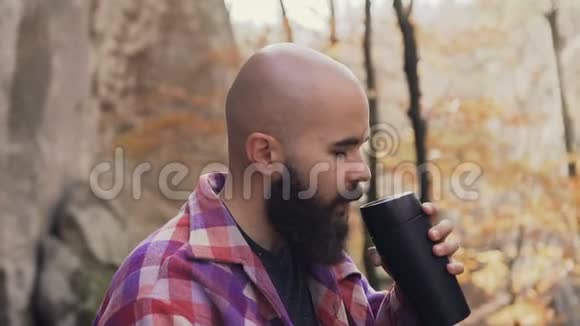 美丽的大胡子男人坐下来在岩石附近的树林里休息手里拿着一个热水瓶喝着茶视频的预览图