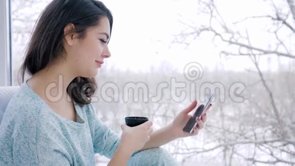 家庭舒适手里拿着小玩意的年轻女子喝着咖啡坐在室内的大窗户附近视频的预览图