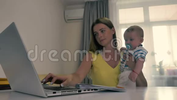 年轻的母亲在家工作手里拿着笔记本电脑抱着孩子视频的预览图