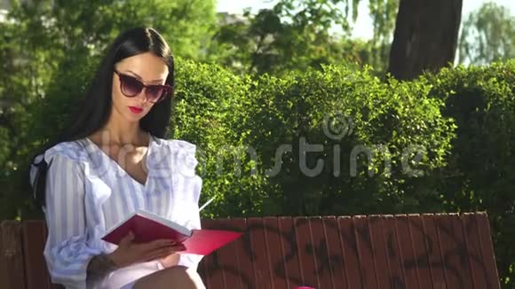 戴墨镜的时髦黑发女人在公园的长椅上看书视频的预览图