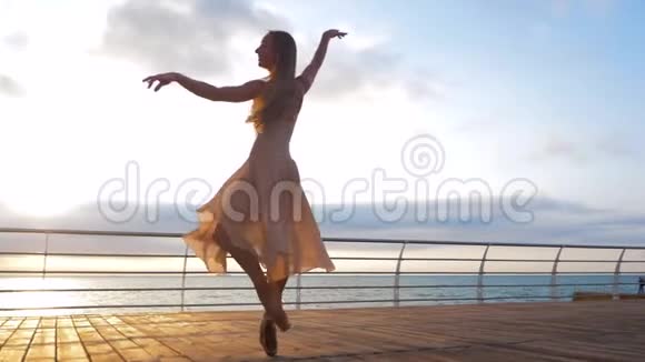 日出时穿着米黄色丝质连衣裙和海堤上的尖角跳舞年轻漂亮的金发女郎视频的预览图