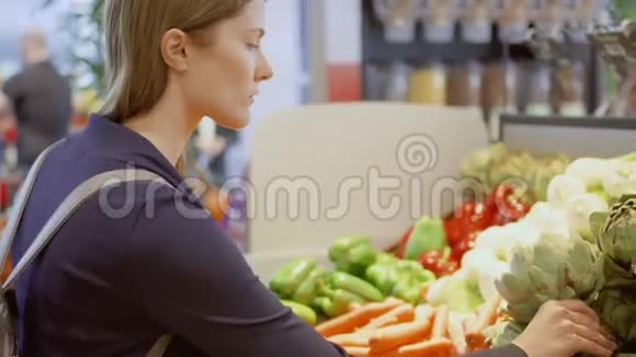 年轻女子在杂货店购物站在蔬菜部门选择新鲜洋蓟视频的预览图